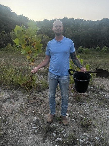 A man on quarry gravel at dusk, a young sycamore in one hand, a bucket and shovel in the other.