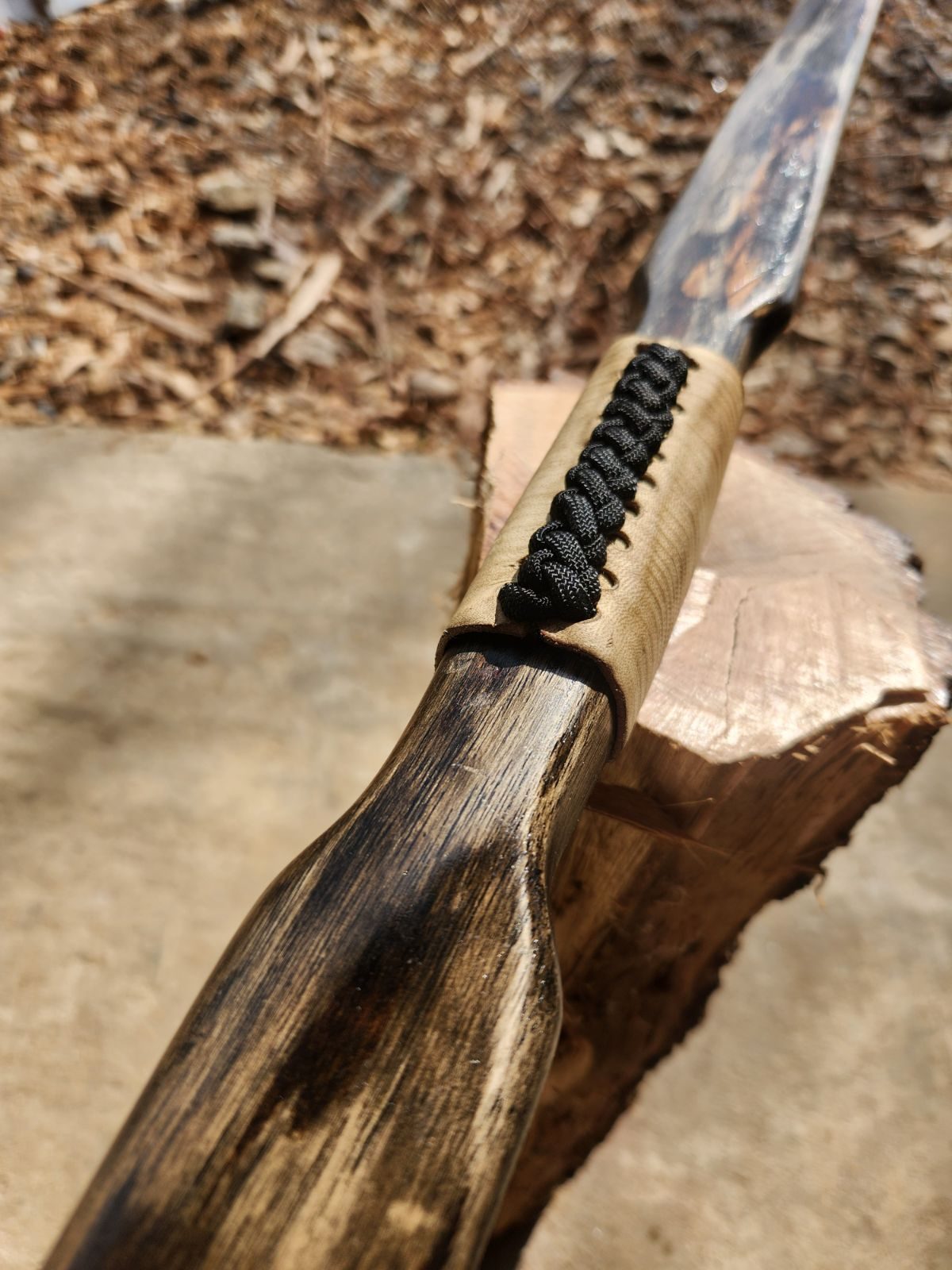 A handmade bow shaped from bodark, dark grain running the length of the limb, its grip wrapped in black cord, resting on a fresh-cut log.